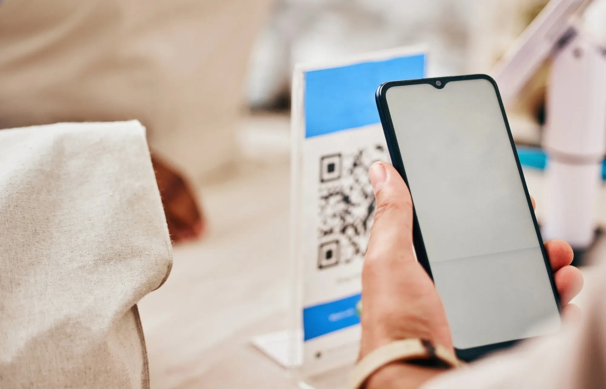 Eine Hand hält ein Smartphone vor einen Aufsteller mit QR-Code, der auf einem Ladentresen steht